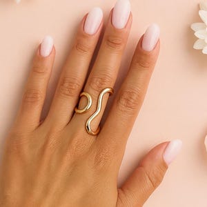 May include: A hand adorned with a gold ring and bracelets, set against a pink backdrop with white flowers and petals. The ring features a distinctive, flowing design. The bracelets are made of gold beads.