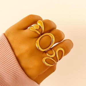 May include: Four gold rings with abstract designs are displayed on a hand. The rings feature shapes like a heart, an oval, and curved lines. The background is a light pink colour.