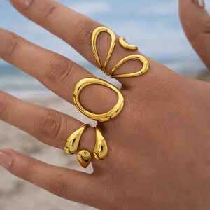 K&ouml;nnte beinhalten: Drei goldfarbene Ringe mit glatten, polierten Oberfl&auml;chen. Der obere Ring hat eine offene, l&auml;ngliche Tropfenform. Der mittlere Ring ist ein gro&szlig;er, abgerundeter ovaler Bandring. Der untere Ring besteht aus zwei tropfenf&ouml;rmigen Elementen.