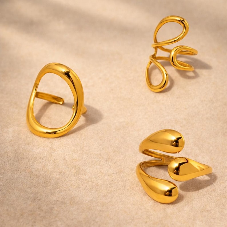 K&ouml;nnte beinhalten: Drei goldfarbene Ringe mit glatten, polierten Oberfl&auml;chen. Ein Ring hat eine gro&szlig;e, offene, unregelm&auml;&szlig;ige Kreisform. Ein weiterer Ring hat ein mehrschichtiges, geschlungenes Design. Der dritte Ring besteht aus zwei &uuml;berlappenden Tropfen.