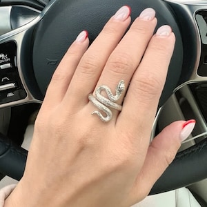May include: A silver snake ring with a textured finish. The ring is worn on the middle finger of a hand with red-tipped nails.