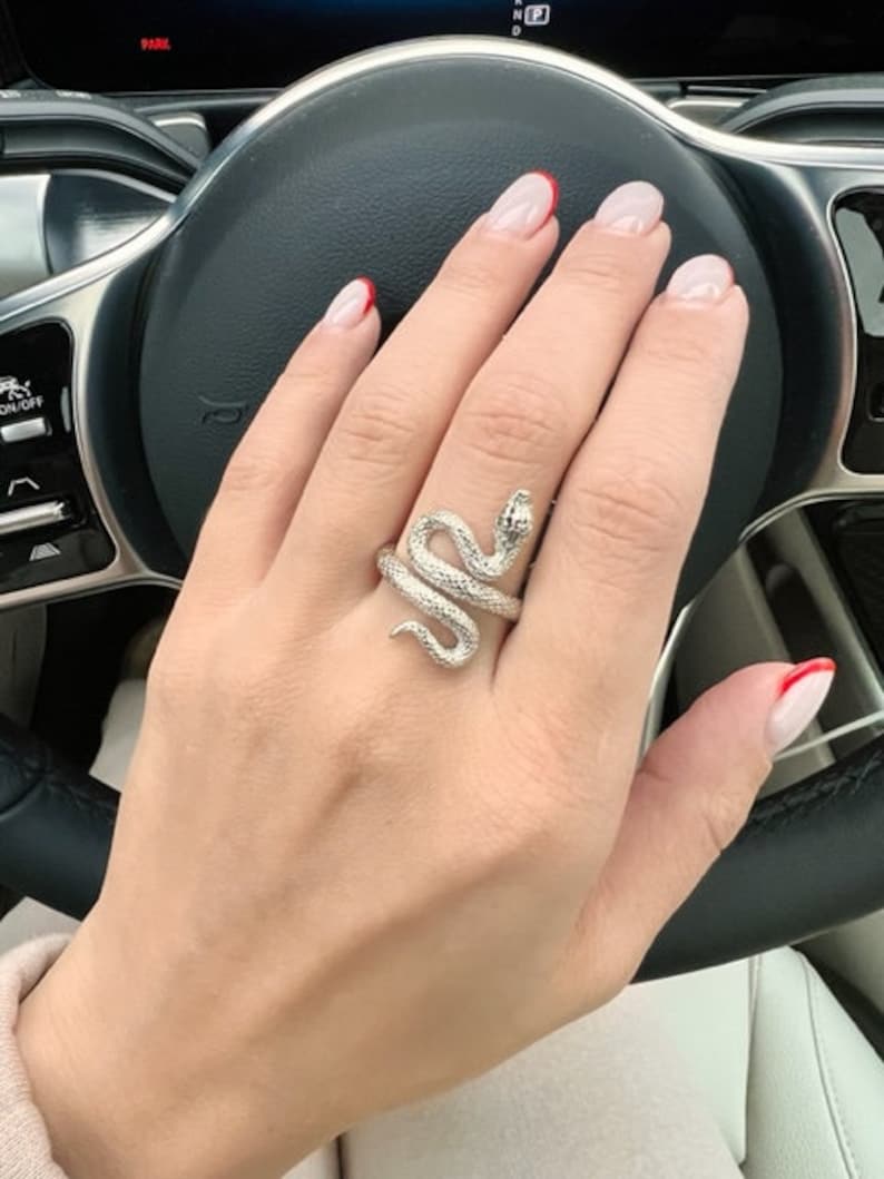May include: A silver snake ring with a textured finish. The ring is worn on the middle finger of a hand with red-tipped nails.