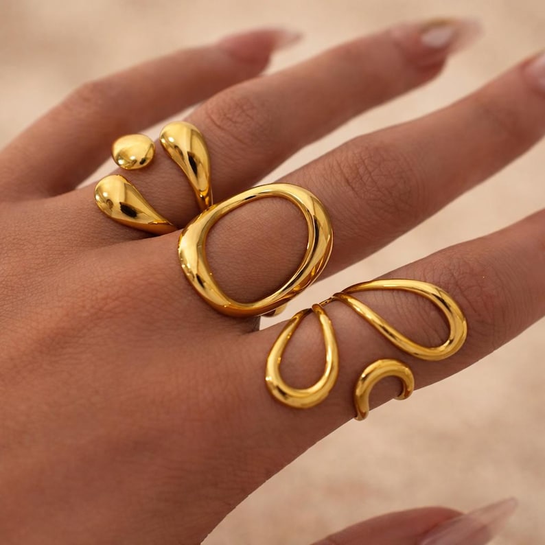 K&ouml;nnte beinhalten: Drei goldfarbene Ringe mit glatten, polierten Oberfl&auml;chen. Ein Ring hat eine gro&szlig;e, offene ovale Form, w&auml;hrend die anderen beiden Ringe asymmetrische, flie&szlig;ende, tropfenartige Ausschnitte und abgerundete Metallformen aufweisen.