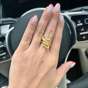 May include: A gold snake ring with a textured finish. The ring is worn on the middle finger of a hand with white nail polish and red tips.