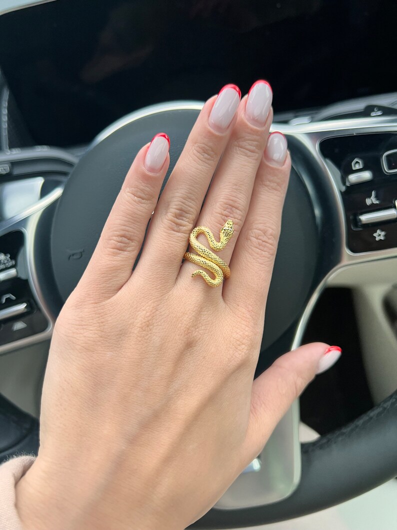 May include: A gold snake ring with a textured finish. The ring is worn on the middle finger of a hand with white nail polish and red tips.