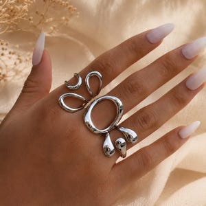 K&ouml;nnte beinhalten: Ein silberfarbener Ring mit einem abstrakten Design aus mehreren Schlaufen, einer gro&szlig;en ovalen Form in der Mitte und kleineren, flie&szlig;enden, organisch wirkenden geschwungenen Formen, die sich &uuml;ber den Finger erstrecken, mit polierter Oberfl&auml;che.