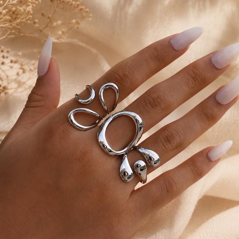 K&ouml;nnte beinhalten: Ein silberfarbener Ring mit einem abstrakten Design aus mehreren Schlaufen, einer gro&szlig;en ovalen Form in der Mitte und kleineren, flie&szlig;enden, organisch wirkenden geschwungenen Formen, die sich &uuml;ber den Finger erstrecken, mit polierter Oberfl&auml;che.