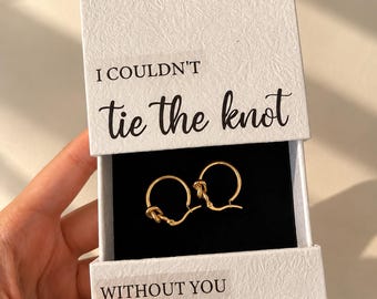 May include: A pair of gold-toned hoop earrings featuring a knot design, presented in a white textured gift box. The box lid is printed with the text I COULDN'T tie the knot, and the base features the text WITHOUT YOU.