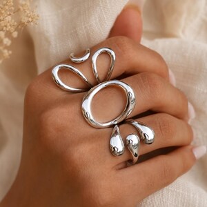 K&ouml;nnte beinhalten: Ein Set aus silberfarbenen Ringen mit organischen, flie&szlig;enden Formen. Das Design umfasst einen gro&szlig;en ovalen Ring in der Mitte, kleinere geschwungene offene Ringe und tropfenf&ouml;rmige Akzente, die den Finger hinunterzulaufen scheinen, alle poliert.