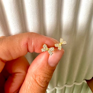 May include: A pair of gold-colored bow-shaped stud earrings. Each earring is encrusted with small, clear gemstones, creating a sparkling effect. The earrings are held between fingers against a neutral background.