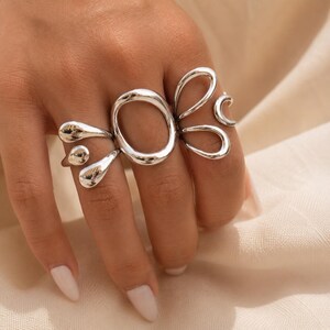 K&ouml;nnte beinhalten: Ein silberfarbener Ring mit einem offenen, unregelm&auml;&szlig;igen ovalen Mittelst&uuml;ck, das von kleineren, glatten, abgerundeten organischen Formen flankiert wird, die sich &uuml;ber den Finger erstrecken. Das Metall hat eine polierte, reflektierende Oberfl&auml;che.