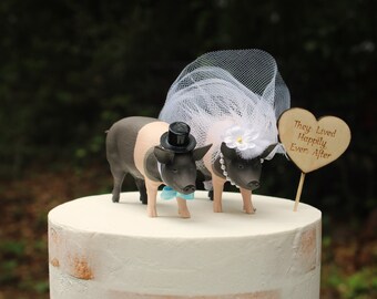 Può includere: Un topper per torta nuziale con due figurine di maiali neri e rosa, uno con un cappello a cilindro e un papillon, l'altro con un velo e perle. Il topper include un cartello di legno a forma di cuore che dice "Vissero felici e contenti".