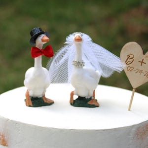 May include: A whimsical wedding cake topper featuring two goose figurines. One goose wears a black top hat and red bow tie, the other a veil and ring. A wooden heart with initials and a date completes the design.