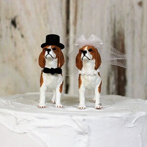 May include: A wedding cake topper featuring a pair of beagle figurines. The groom beagle wears a black top hat and bow tie, while the bride beagle wears a white veil and pearl necklace.