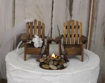 Könnte beinhalten: Ein Hochzeitstortenaufsatz mit zwei Adirondack-Stühlen aus Holz mit einem kleinen Lagerfeuer dazwischen. Die Stühle sind mit weißen Blumen verziert und jeder Stuhl hat ein Gewehr, das auf der Rückenlehne liegt.