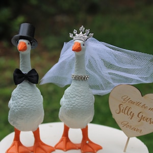 May include: Two white goose figurines with orange beaks and feet. One wears a black top hat and bow tie. The other wears a silver crown, a white tulle veil, and a rhinestone collar. A wooden heart sign reads: You'll be my Silly Goose forever.