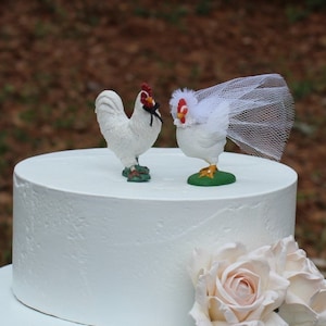 May include: A white wedding cake topper featuring a white rooster wearing a black bow tie and a white hen wearing a white veil. The cake topper is on a white cake with a pink rose.