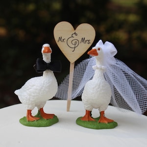 May include: Wedding cake topper featuring two white goose figurines. One goose wears a black bow tie, the other a white veil. A wooden heart-shaped sign with "Mr. & Mrs." is between them. The geese stand on green bases.