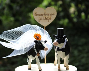 Op de afbeelding: Een bruidstaarttopper met een zwart-witte koe met een sluier en een zonnebloem, en een zwart-witte koe met een hoge hoed en vlinderdas. Het bordje staat "Gonna love you for Heifer."