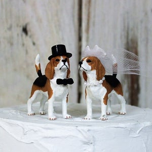 May include: A wedding cake topper featuring a pair of beagle figurines. The groom beagle wears a black top hat and bow tie, while the bride beagle wears a white veil and pearl necklace.