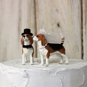 May include: A wedding cake topper featuring a pair of beagle figurines. The groom beagle wears a black top hat and bow tie, while the bride beagle wears a white veil and pearl necklace.
