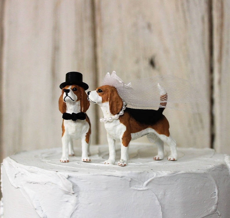 May include: A wedding cake topper featuring a pair of beagle figurines. The groom beagle wears a black top hat and bow tie, while the bride beagle wears a white veil and pearl necklace.