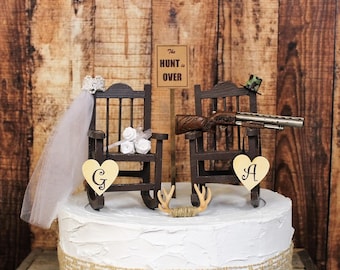 Könnte beinhalten: Hochzeitstortenaufsatz mit zwei Miniatur-Schaukelstühlen aus Holz. Ein Stuhl hat einen Schleier und Blumen, der andere eine Schrotflinte und einen Camouflage-Hut. Ein Schild liest "The Hunt is Over". Der Kuchen ist mit einem Herz mit dem Buchstaben "G" und einem weiteren mit dem Buchstaben "A" verziert.