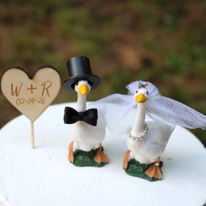 May include: Two white goose figurines as a cake topper. One goose wears a black top hat and bow tie, the other wears a white tulle veil and a rhinestone necklace. A wooden heart-shaped sign on a stick displays the text W + R and 02-14-26.