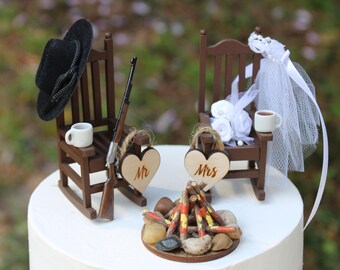 Könnte beinhalten: Hochzeitstortenaufsatz mit Miniatur-Holzstühlen, einem Lagerfeuer und herzförmigen Schildern mit der Aufschrift "Mr" und "Mrs". Ein schwarzer Hut und ein Gewehr liegen auf einem Stuhl, der andere hat einen Schleier und Blumen. Auf jedem Stuhl steht eine Tasse Kaffee.