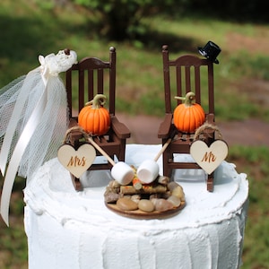 Puede incluir: Un adorno para pastel de boda con dos mecedoras marrones con calabazas y un letrero en forma de corazón que dice "Mr." y "Mrs.". Las sillas están colocadas alrededor de una fogata en miniatura con malvaviscos.