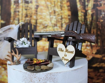 Könnte beinhalten: Ein Hochzeitstortenaufsatz mit zwei Holzstühlen, einer Schrotflinte, einem herzförmigen Schild mit der Aufschrift "Die Jagd ist vorbei", einem kleinen Holzstapel und einem Geweih.
