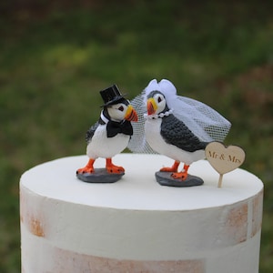 May include: A wedding cake topper with two puffins. One is dressed in a top hat and bow tie, the other in a veil. They stand on a white cake, with a wooden heart that says "Mr & Mrs".