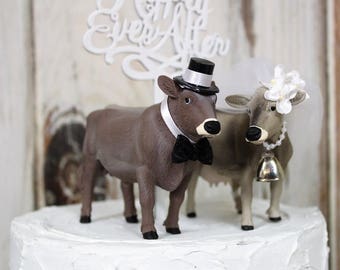 Op de afbeelding: Een bruidstaarttopper met een bruine koe die een zwarte hoed en vlinderdas draagt en een witte koe die een witte bloemenkrans draagt. De taarttopper staat op een witte taart met de woorden "Happily Ever After" op de taart.