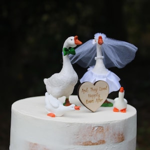 May include: A wedding cake topped with goose figurines. One goose wears a green bow tie, another a veil and dress. A small wooden heart with the text "And They Lived Happily Ever After" is also on the cake.