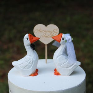 May include: Two white carved wooden goose figurines with orange beaks and feet. One goose wears a black bow tie, and the other wears a white veil with a small flower. A wooden heart-shaped sign on a stick stands between them, inscribed with the text Anthony & Julia.