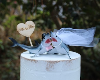 May include: A whimsical wedding cake topper featuring two shark figurines. One shark wears a top hat and pink bow tie, the other a veil and pearl necklace. A wooden heart with "Mr & Mrs" is also included.