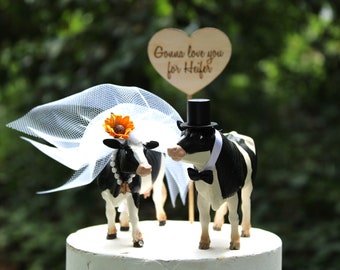 Op de afbeelding: Een bruidstaarttopper met een zwart-witte koe die een witte sluier en een zonnebloem draagt, en een zwart-witte koe die een hoge hoed en een strikje draagt. Het bord achter de koeien luidt "Gonna love you for heifer."