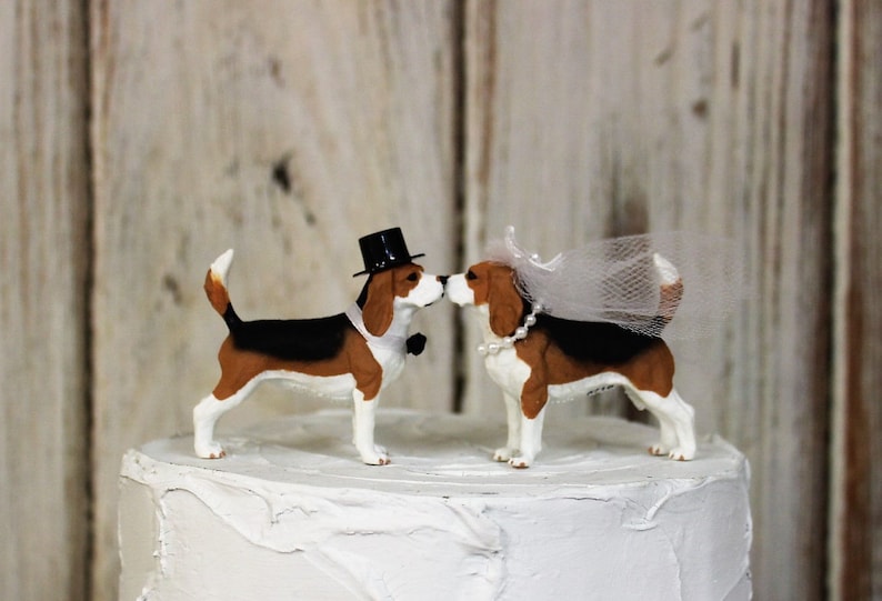 May include: A wedding cake topper featuring two beagle figurines, one wearing a top hat and the other a veil. The figurines are kissing.