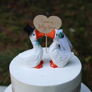May include: Two white carved goose figurines on a white cake. The left goose wears a black top hat and a teal bowtie. The right goose wears a white veil with a teal flower and a pearl necklace. A wooden heart sign reads I found my Silly Goose.