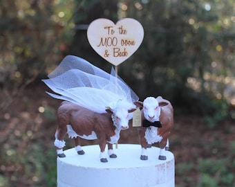 Op de afbeelding: Een bruidstaarttopper met een bruine en witte koe die een witte sluier draagt en een witte en zwarte stier die een zwarte vlinderdas draagt. De topper bevat een houten hartvormig bord met de tekst "To the MOO ooo & Back".