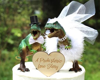 May include: Two green Tyrannosaurus rex dinosaur figurines as a cake topper. One wears a black top hat and white bow tie. The other wears a white tulle veil, a white feather boa, and a pearl necklace. A wooden heart sign reads A Prehistoric Love Story.