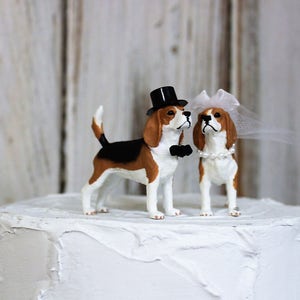 May include: A white wedding cake with a brown and white beagle wearing a black top hat and a bow tie and a white beagle wearing a white veil and a pearl necklace on top.