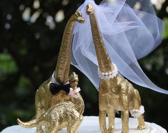 May include: Gold dinosaur wedding cake topper with a bride and groom dinosaur, the bride wearing a white veil and a pearl necklace, and the groom wearing a black bow tie. A smaller dinosaur stands behind the couple.