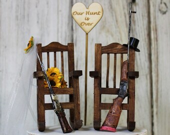 Könnte beinhalten: Ein Hochzeitstortenaufsatz mit zwei Holz-Schaukelstühlen, jeder mit einer Schrotflinte und einem Sonnenblumenstrauß. Die Stühle stehen auf einer weißen Torte mit einem Schild in Form eines Holzh Herzens, auf dem "Unsere Jagd ist vorbei" steht.