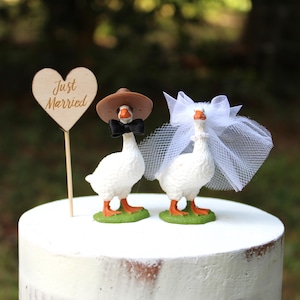 May include: Two white goose figurines standing on green bases. One goose wears a brown hat and black bow tie, the other wears a white tulle veil. A wooden heart-shaped sign on a stick with the text Just Married stands beside them.