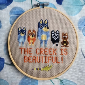 Könnte beinhalten: Ein Kreuzstich-Stickrahmen mit fünf Zeichentrickfiguren in Blau, Orange, Schwarz und Braun. Der Text "THE CREEK IS BEAUTIFUL!" ist mit orangefarbenem Faden gestickt. Am unteren Rand ist eine kleine grüne und gelbe Kreatur gestickt.
