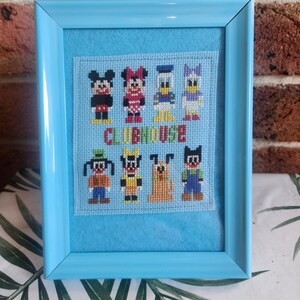 Op de afbeelding: Een ingelijste kruissteek met stripfiguren, waaronder Mickey Mouse, Minnie Mouse, Donald Duck en Daisy Duck. Het woord "CLUBHOUSE" is onder de figuren gestikt. De lijst is lichtblauw.