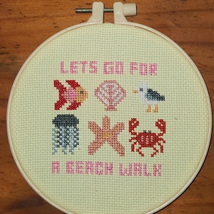 May include: A cross-stitch embroidery hoop with a light yellow background. The text "LET'S GO FOR A BEACH WALK" is stitched in pink. The design includes a fish, seashell, seagull, jellyfish, starfish, and crab.