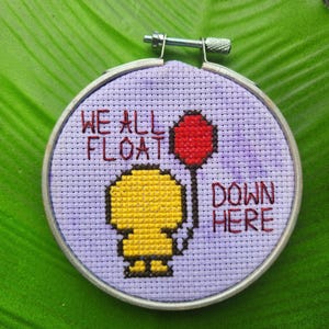 Puede incluir: Un bastidor de bordado de punto de cruz con una figura amarilla sosteniendo un globo rojo. El texto "WE ALL FLOAT DOWN HERE" está bordado en rojo sobre un fondo lavanda. El bastidor se asegura con un cierre metálico.