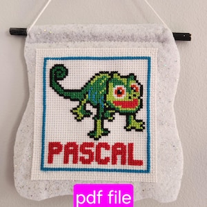 Puede incluir: Una pancarta de fieltro blanco con un borde azul y una imagen de punto de cruz de Pascal, el camaleón de Enredados de Disney, con la palabra "PASCAL" en letras rojas debajo de la imagen. La pancarta está colgada de una cuerda blanca y tiene una varilla de madera en la parte superior.
